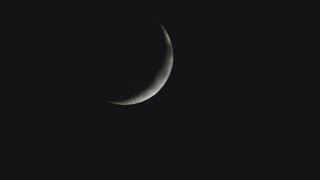 Heat waves dance over the edge of a crescent moon and a few thin clouds pass in front of it as it slowly sinks lower in the dark night sky.