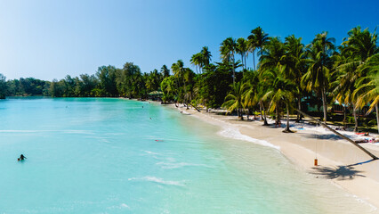 Clear turquoise waters gently lap against the soft white sand of a serene beach in Koh Kood. Lush palm trees sway as visitors relax and enjoy the tropical paradise during a sunny day.