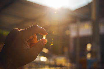 Image of a hand holding a shiny yellow vitamin pill against a sunny orange-golden sky in the morning and evening.