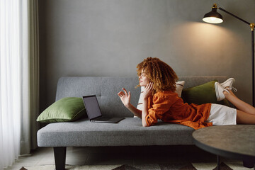 A young woman of African descent sits on a modern gray sofa, thoughtfully using a laptop and...