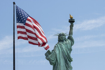 Back view of the Statue of Liberty on the Liberty Island in New York, with a waving American flag by the side, against a blue sky and clouds.