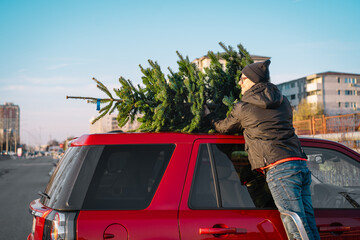 Man secures a Christmas tree on top of a red car in an urban setting.