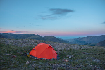 Orange tent on grassy stony pass with view to alpine valley and mountain range silhouette far away under clouds in sunset color sky. Red tent in high mountains under lenticular clouds in sunset sky.
