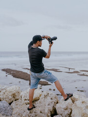 Full body asian male photographer taking photos while standing on the coral rocks by the beach on the island of Bali, Indonesia. Travel, leisure and vacation concept.