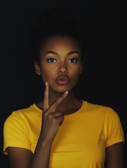 Fototapeta premium Stupefied secret dark skinned woman makes gesture quietly, asks remain silent, gossips about something, looks mysteriously as tells secret, wears bright yellow t shirt, stands indoor. Shh, dont speak