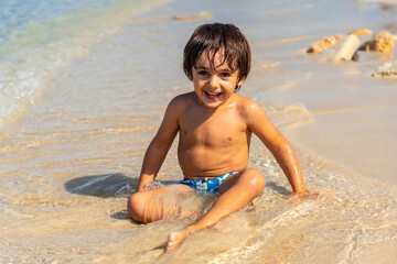 A young boy is sitting on the beach, smiling and enjoying the water