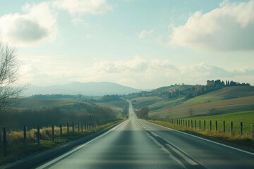 A long, straight road traverses rolling green hills, bordered by a simple fence. Soft clouds float in a blue sky, creating a peaceful atmosphere in the countryside.