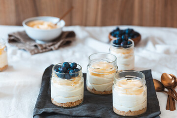 Glass jars of layered lemon cheesecake topped with lemon curd and fresh blueberries sit on a dark cloth. A bowl of blueberries and another with lemon curd are seen in the background.