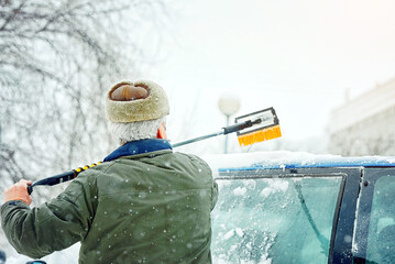 Man sweeps snow off the rood of car during heavy snowfall in an icy parking area. Senior man cleans...