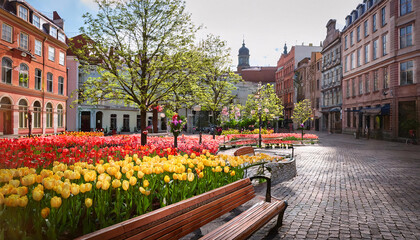 Obraz premium A lively city square adorned with vibrant red and yellow tulips, inviting benches, and blossoming trees under a bright spring sun create a cheerful atmosphere ideal for relaxation