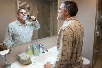 Senior man brushing teeth with electric toothbrush in bathroom mirror