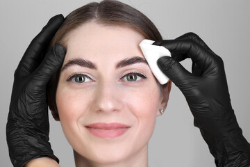 Brow lamination. Cosmetologist wiping woman's eyebrows with cotton pad against grey background,...