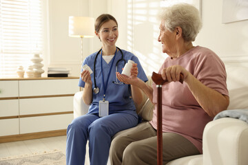 Caregiver giving pills to senior woman on sofa indoors. Home health care service
