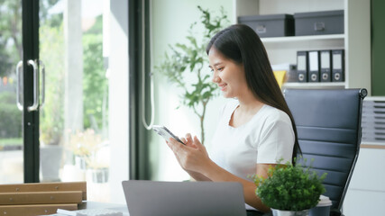 An Asian businesswoman works at her desk in a modern office, smiling while using a tablet to review financial analysis reports, manage accounting tasks, and oversee banking and loan documentation