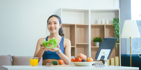 A happy Asian woman in sportswear sits on a sofa at a table, enjoying a salad bowl, fruits, fresh vegetables, orange juice, and energy supplements after a home fitness workout