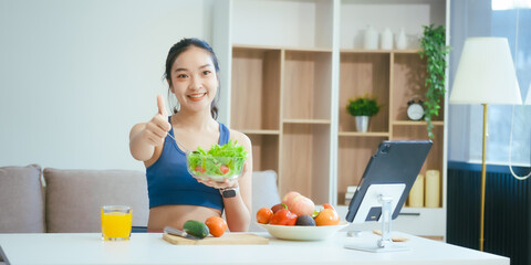 A happy Asian woman in sportswear sits on a sofa at a table, enjoying a salad bowl, fruits, fresh vegetables, orange juice, and energy supplements after a home fitness workout