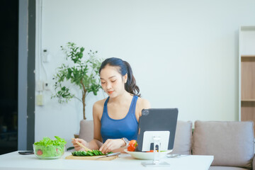 A happy Asian woman in sportswear sits on a sofa at a table, enjoying a salad bowl, fruits, fresh vegetables, orange juice, and energy supplements after a home fitness workout