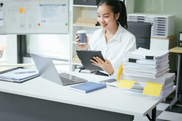 An Asian businesswoman sits at her desk in the office, working on her laptop, checking documents, and planning business strategies to enhance productivity and achieve professional goals