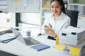 An Asian businesswoman sits at her desk in the office, working on her laptop, checking documents, and planning business strategies to enhance productivity and achieve professional goals