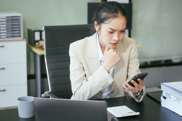Obraz premium An Asian businesswoman sits at her office desk, smiling happily while working on her laptop. She reviews documents, plans new business strategies, and manages tasks efficiently to achieve success