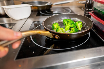 A hand holds a frying pan with steaming broccoli on an induction stove. The kitchen setting features various kitchenware, showcasing a vibrant cooking scene.