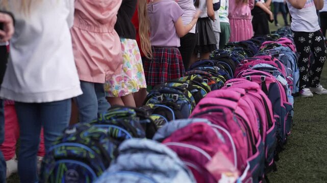 Children gathered on a school field, standing in a row near a long line of colorful backpacks, symbolizing education and community. Concept of back-to-school preparation and school events