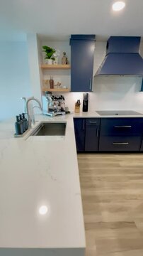 Vertical video of a modern coastal kitchen in a beachy luxury condo. The newly built custom kitchen features dark blue cabinets, white waterfall island, white countertops, and oak wood accents.