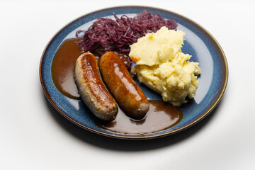 , Deutschland, 29.12.2024,  Bratwürste mit dunkler Soße, Rotkohl und cremigem Kartoffelpüree auf einem blauen Teller – ein klassisches deutsches Gericht.