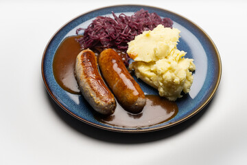 , Deutschland, 29.12.2024,  Bratwürste mit dunkler Soße, Rotkohl und cremigem Kartoffelpüree auf einem blauen Teller – ein klassisches deutsches Gericht.