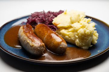 , Deutschland, 29.12.2024,  Bratwürste mit dunkler Soße, Rotkohl und cremigem Kartoffelpüree auf einem blauen Teller – ein klassisches deutsches Gericht.