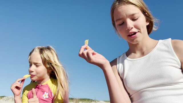 Two sister girls, eight and twelve years old, are sitting on the beach of the Atlantic Ocean, eating a snack, and enjoying the sun and summer.