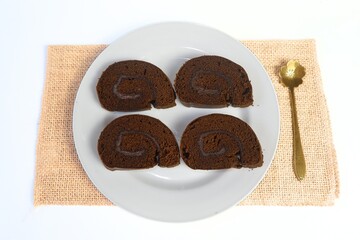slice of chocolate swiss roll cake or bolu gulung coklat made with sponge cake base, layer of butter cream, then rolled into roll cake. Served on white plate.