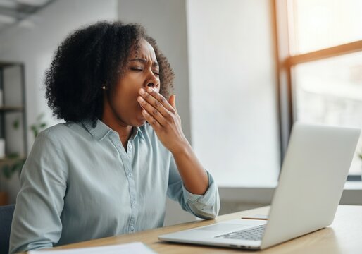 Exhausted office worker covering mouth while yawning at desk in front of laptop