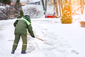 Naklejka premium Worker shoveling heavy snow in city park, seasonal maintenance in snowy and icy conditions. Utility worker cleaning snow from sidewalk with shovel in cold winter weather after heavy snowstorm