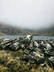 A view of a lake surrounded by mountains, shrouded in mist. The foreground features rock formations covered in green moss and short grass.