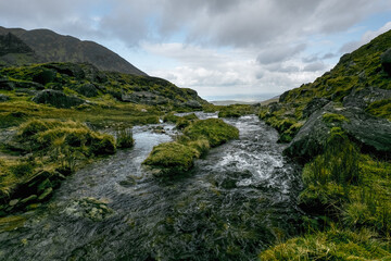 Rocky terrain surrounds a flowing stream. Vegetation thrives along the water's edge. A distant view is visible.