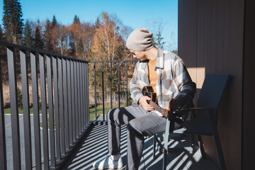 Young musician sits on a sunny balcony, immersed in playing the ukulele. Surrounded by autumn...