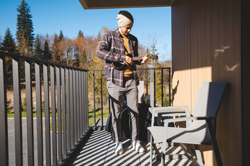 young musician stands on a sunlit balcony, playing the ukulele while gazing into the distance....