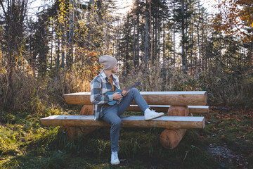 Fototapeta premium Musician sits on a rustic wooden bench in a sunlit forest, playing the ukulele. Surrounded by autumn foliage and warm sunlight streaming through the trees