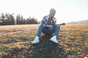 Young man sits on a grassy field in the warm autumn sunlight, smiling as he plays the ukulele. Surrounded by golden tones and distant trees, the moment reflects joy, creativity, and a deep connection