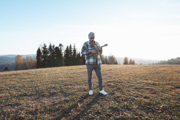 Musician stands in an open field during a sunny autumn day, holding a ukulele. Surrounded by hills and trees, the serene scene reflects freedom, creativity, and a connection to nature