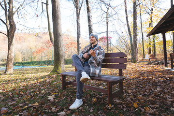 Fototapeta premium Enthusiast sits on a wooden bench in a park, playing the ukulele amidst autumn leaves. Surrounded by trees and bathed in soft sunlight, he enjoys a tranquil moment filled with creativity