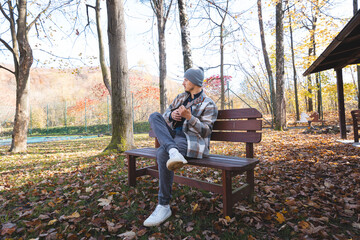 Fototapeta premium Enthusiast sits on a wooden bench in a park, playing the ukulele amidst autumn leaves. Surrounded by trees and bathed in soft sunlight, he enjoys a tranquil moment filled with creativity