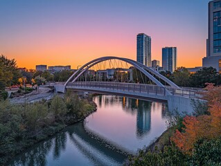 Naklejka premium Sunset view of modern arched bridge over calm river with cityscape background.