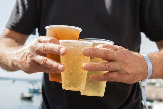 The photograph features hands holding four plastic cups of different beverages in an outdoor setting.