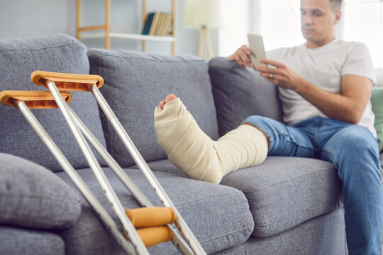 Handicapped young man sitting at home on sofa in rehabilitation with crutches using mobile phone. Male person with injured leg after accident in plaster or splint with walking stick.