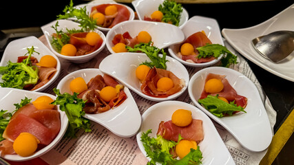 Elegant appetizers featuring prosciutto, melon balls, and greens in white bowls on a table, perfect for gourmet holiday catering