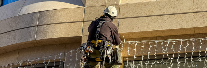 A construction worker installs Christmas lights on a building facade, emphasizing safety and...