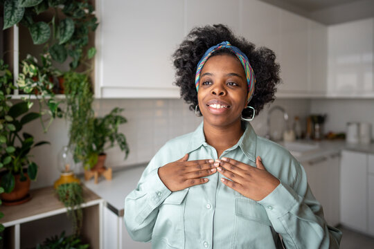 Joyful African American woman at home with hands over heart feels gratitude, inner peace. Self-care, emotional wellbeing, mindfulness. Serene black female smiling warmly by self-love, faith, optimism