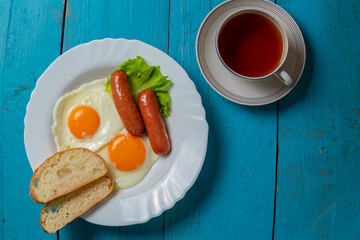 Breakfast fried eggs with sausages fresh toast and tea in a cup on a blue wooden table